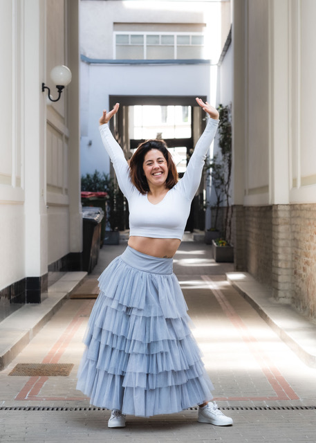 Yoga Lehrerin Joana mit weißem Shirt und hellblauem langen Rock mit breitem Lächeln und hochgestreckten Armen